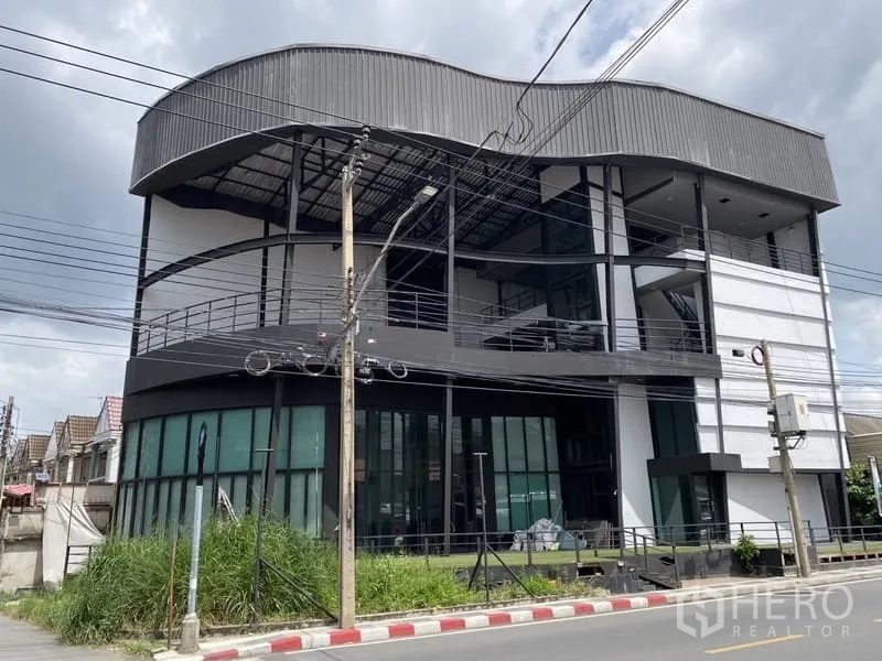 Showroom for rent in Lat Phrao, Bangkok - Four-storey showroom building in Lat Phrao with curved metal roof and full-height glass frontage on a street corner.