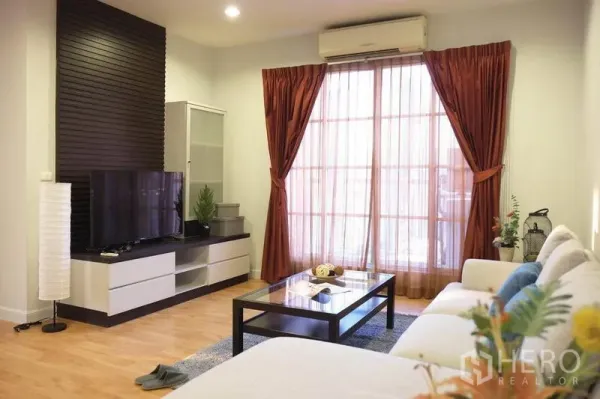 Condominium for rent in Khlong Toei, Bangkok - Bright living room with sectional sofa, TV console, wood floors, and sliding glass doors with rust curtains.