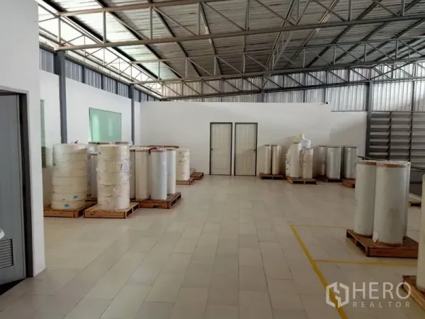 Warehouse for rent in Bang Kapi, Bangkok - Spacious warehouse interior showing steel truss roof, tiled floor, white partition walls and pallets of rolled materials.