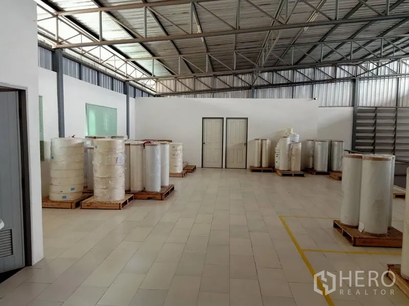 Warehouse for rent in Bang Kapi, Bangkok - Spacious warehouse interior showing steel truss roof, tiled floor, white partition walls and pallets of rolled materials.