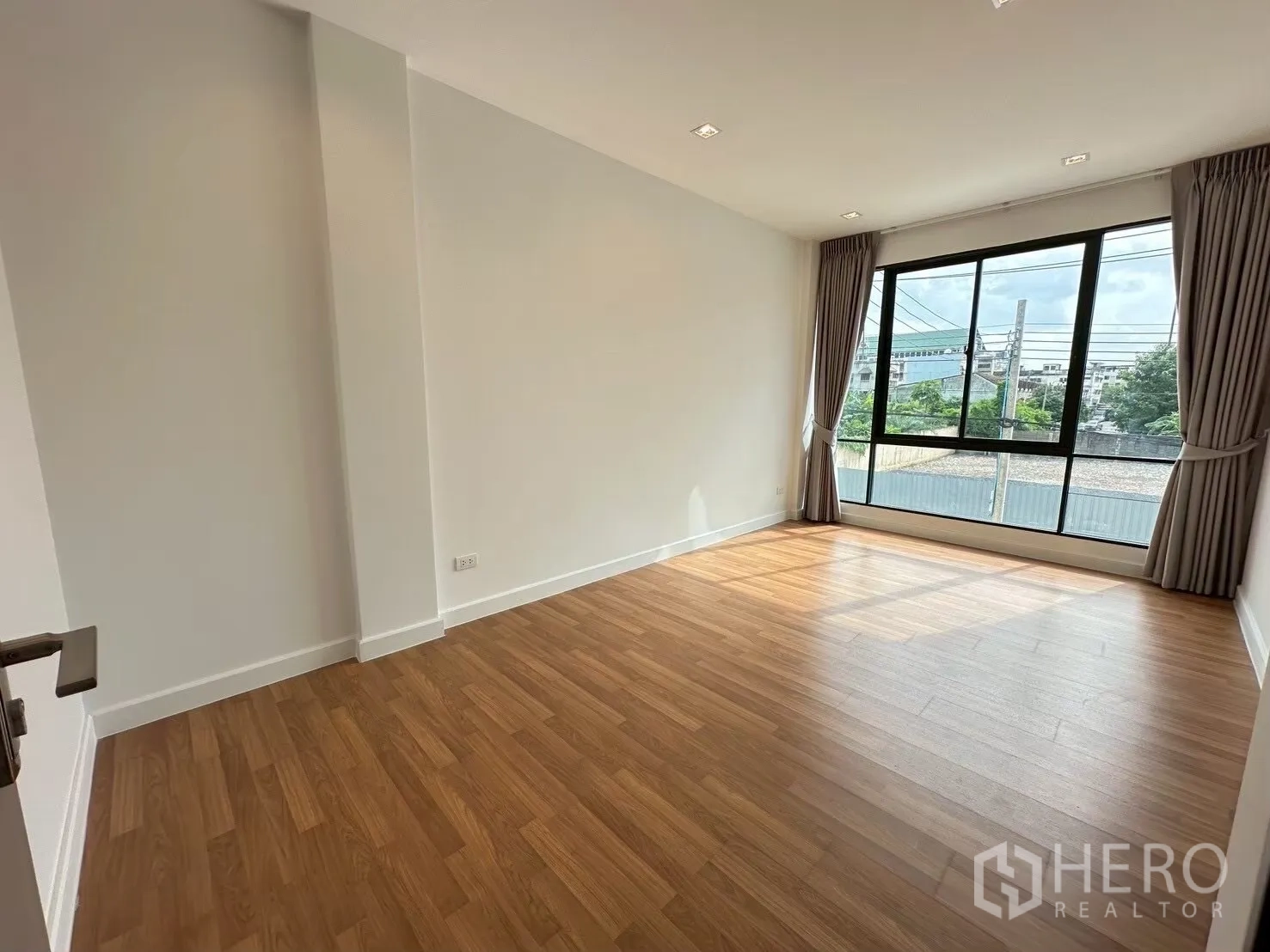Home Office for rent in Lat Phrao, Bangkok - Sunlit bedroom with wide windows, wood flooring, and curtain set overlooking the street.