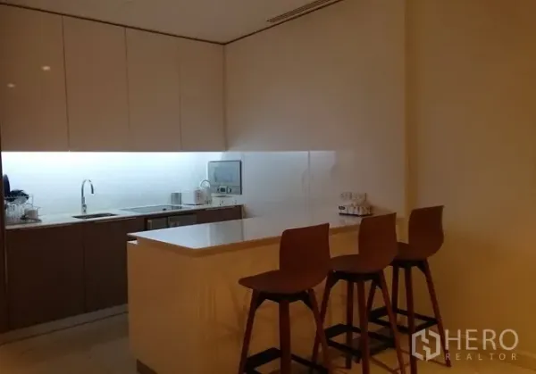 Condominium for rent in Pathum Wan, Bangkok - Modern kitchen with island breakfast bar, three wooden stools, sleek cabinetry and under-cabinet lighting.