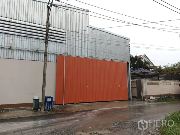 货栈 出租在Khlong Sam Wa, Bangkok - 街景视角的仓库立面，橙色大门与镀锌板外墙，旁侧为住宅围墙。