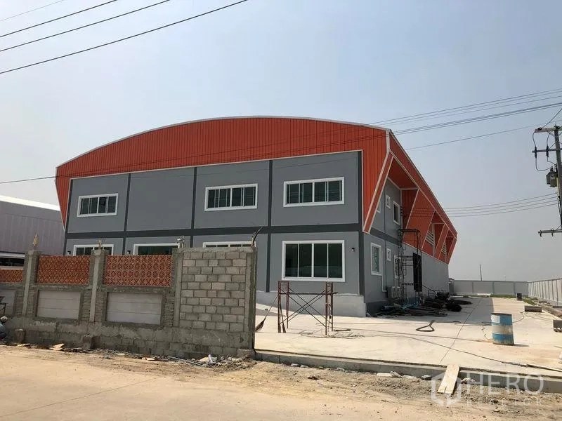 Factory for sale in Mueang Samut Sakhon, Samut Sakhon - Side view of a new industrial factory with orange cladding, wide windows and a spacious concrete yard with utility lines.