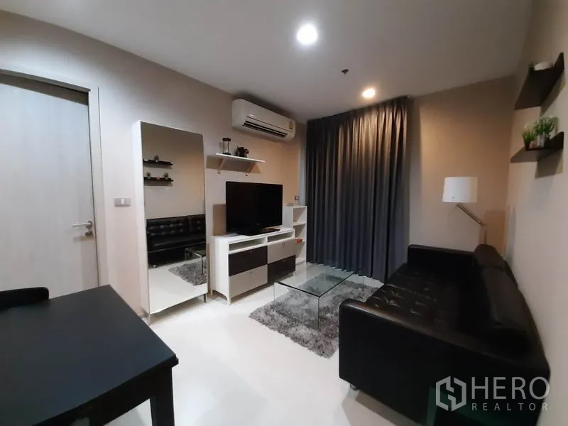 Condominium for rent or sale in Khlong Toei, Bangkok - Cozy living room with black leather sofa, TV console, large mirror and glass coffee table beneath grey blackout curtains.