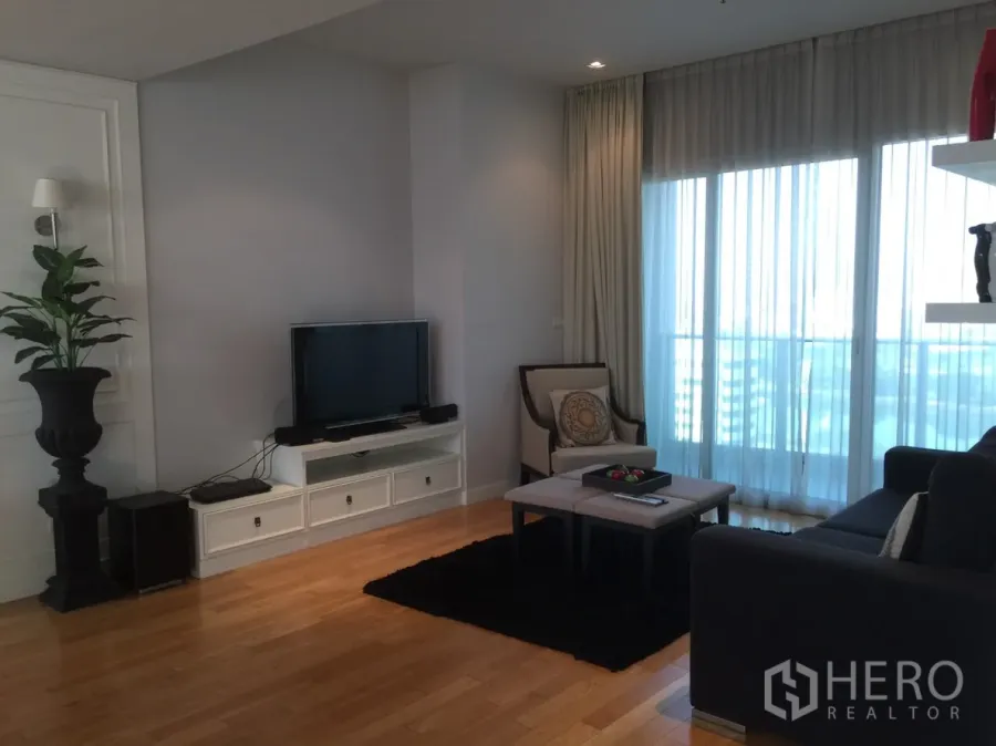 Condominium for rent in Khlong Toei, Bangkok - Cozy living room with TV console, armchair and sliding glass doors to a balcony with sheer curtains.