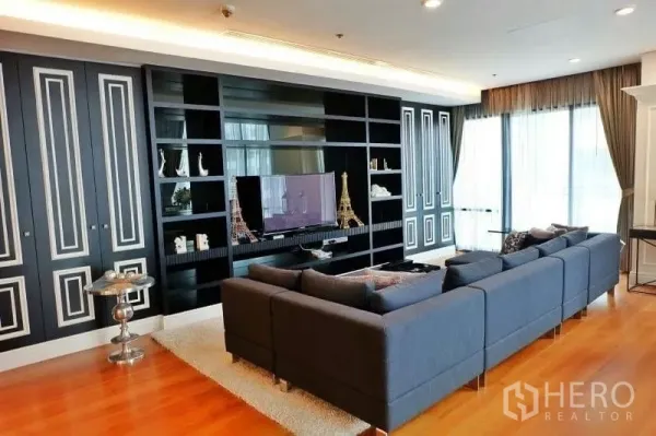 Condominium for rent in Khlong Toei, Bangkok - Spacious living room featuring a dark built-in media wall, large sectional sofa and tall curtained windows.