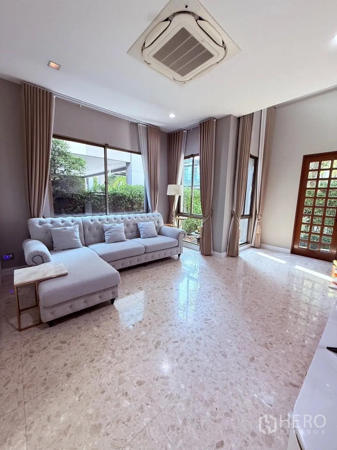 Detached House for rent or sale in Prawet, Bangkok - Bright living room with L-shaped fabric sofa, big windows, tiled floor, and garden views.