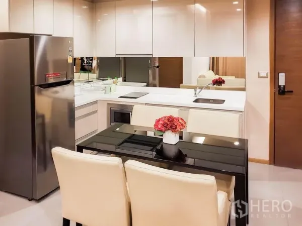 Condominium for rent in Bang Rak, Bangkok - Modern kitchenette with glossy white cabinets, mirrored backsplash, induction hob, stainless refrigerator, and a four-seat dining table.