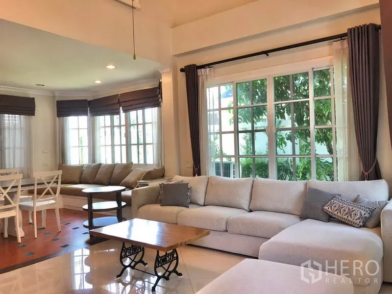 Detached House for rent in Mueang Samut Prakan, Samut Prakan - Bright living room with large L‑shaped sofa, coffee table, wide windows and curtains.
