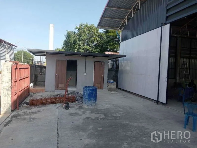 Warehouse for rent in Lam Luk Ka, Pathum Thani - Side yard with small utility rooms, wide access gate and corrugated steel walls.