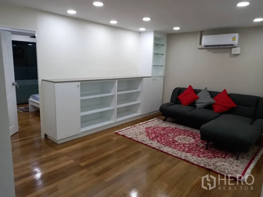 Condominium for rent in Khlong Toei, Bangkok - Cozy living room with built-in white shelves, gray sectional sofa with red cushions, and recessed lighting.