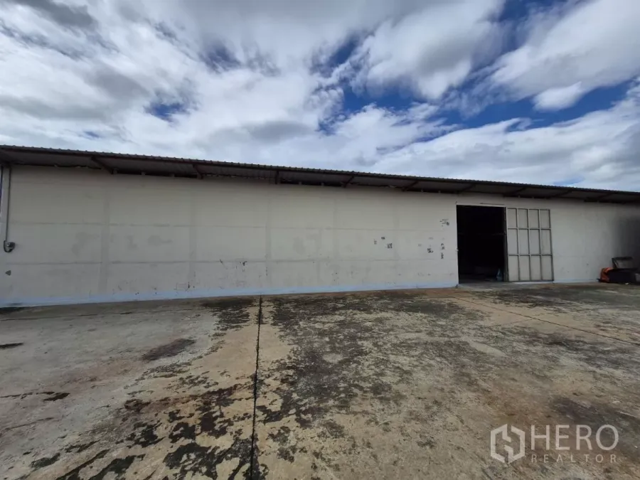 Warehouse for rent in Mueang Pathum Thani, Pathum Thani - Long single-storey warehouse exterior with sliding metal door and wide concrete loading yard under a cloudy sky.