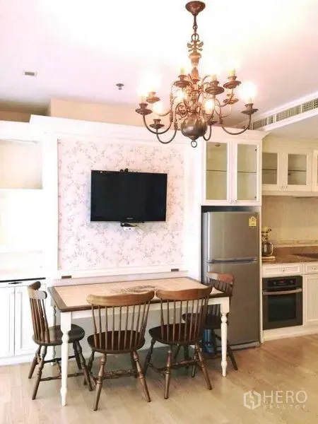 Condominium for rent in Khlong Toei, Bangkok - Dining nook with wooden table and chairs, wall-mounted TV and compact kitchen featuring a stainless fridge and glass-front cabinets.