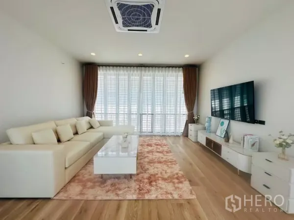 Detached House for rent or sale in Bang Kapi, Bangkok - Bright living room with L-shaped sofa, TV wall, sheer curtains and wood flooring.