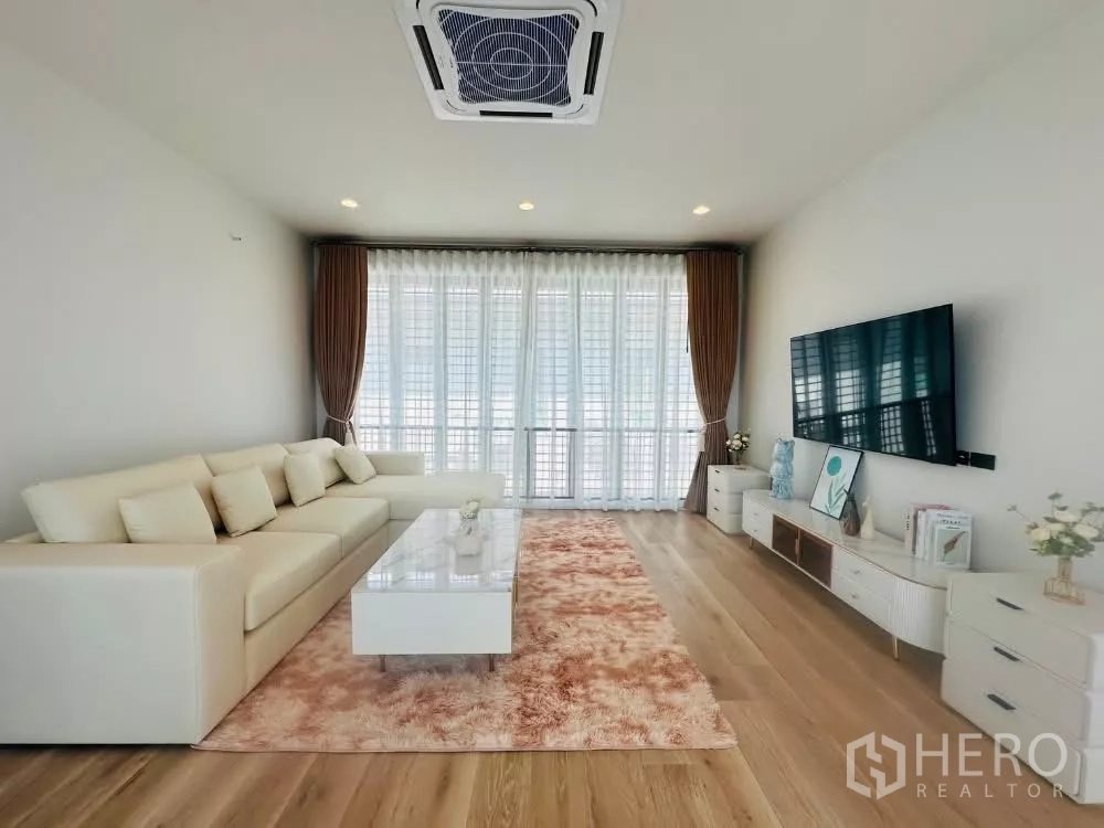 Detached House for rent or sale in Bang Kapi, Bangkok - Bright living room with L-shaped sofa, TV wall, sheer curtains and wood flooring.