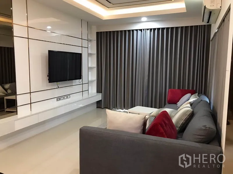 独立式住宅 出租在Bang Phli, Samut Prakan - 客厅明亮，内置电视墙、灰色窗帘、吊顶灯光与L形沙发。