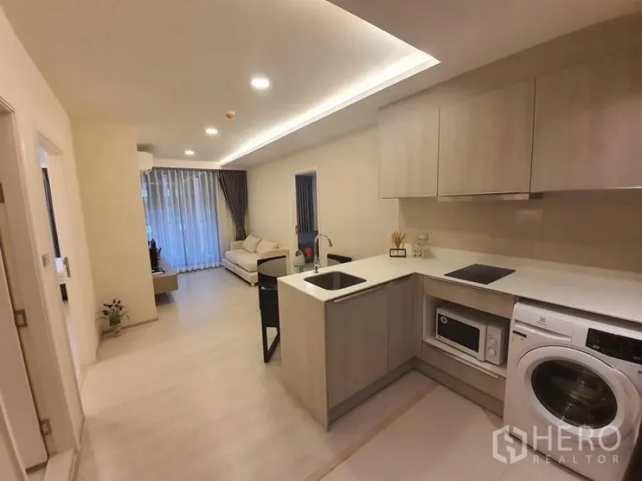 Condominium for rent in Khlong Toei, Bangkok - Open-plan kitchen with built-in cabinets, washer and island facing a bright living room with sofa and full-height curtains.