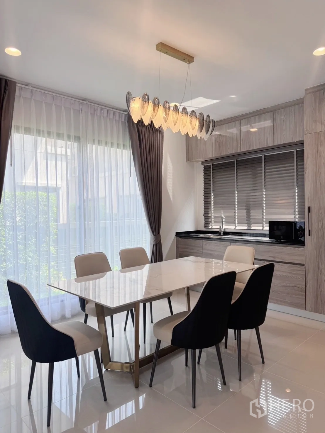 Detached House for rent or sale in Bang Kapi, Bangkok - Dining area with contemporary chandelier next to built-in kitchen cabinetry and window blinds.