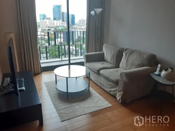 Condominium for rent in Khlong Toei, Bangkok - Bright living room with sofa, round coffee table, TV console and a balcony overlooking Bangkok’s skyline.