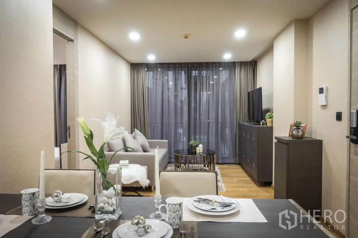 Condominium for rent in Pathum Wan, Bangkok - Open-plan living and dining area with a grey sofa, TV console, and floor-to-ceiling curtains to the balcony.