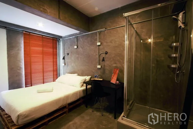 Office Space for rent or sale in Bang Rak, Bangkok - Compact bedroom with concrete walls, a glass shower enclosure, and a work desk.