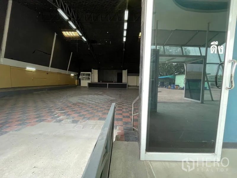 Warehouse for rent in Sam Phran, Nakhon Pathom - View from the glass door into the open storage hall with checkered tile flooring.