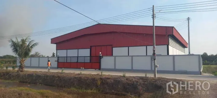 货栈 出租或出售在Khlong Luang, Pathum Thani - 侧立面，红色钢结构仓库与围墙，临铺装道路及电线杆。