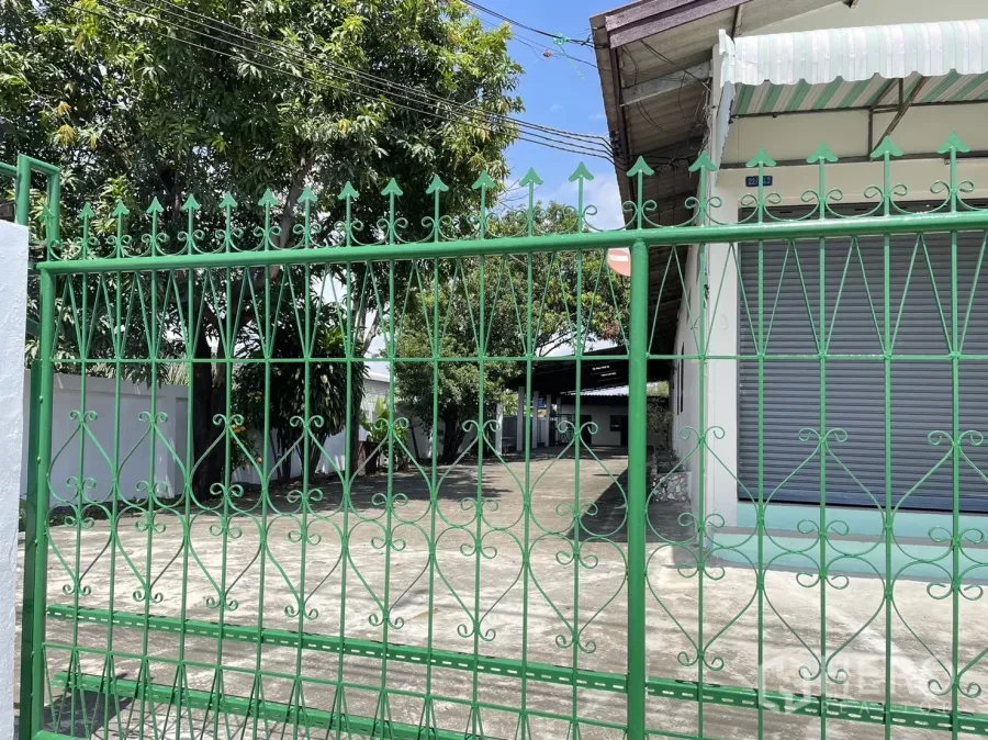 Factory for rent or sale in Pathum Thani - Ornate green gate opening to a concrete driveway with a white factory and roller shutters behind trees.