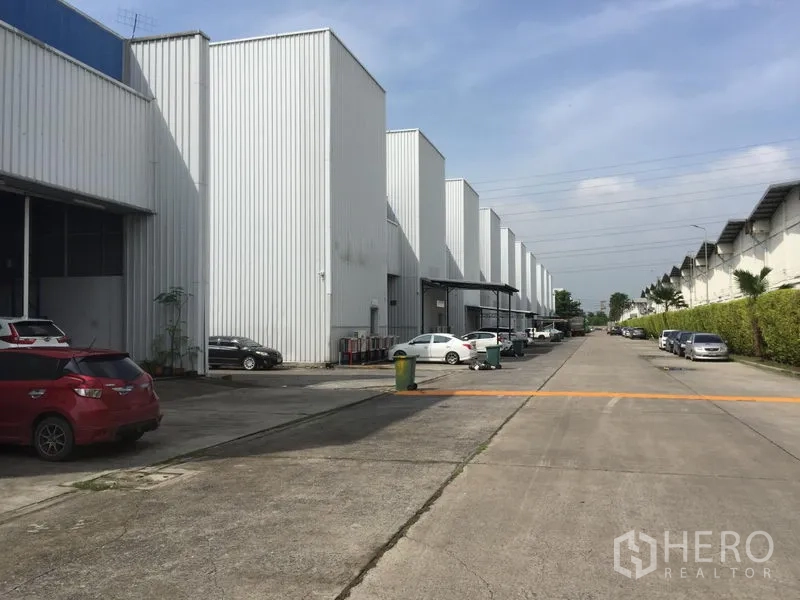 Factory for rent in Khlong Luang, Pathum Thani - Row of warehouse units with high gabled roofs along a wide concrete road inside an industrial complex.