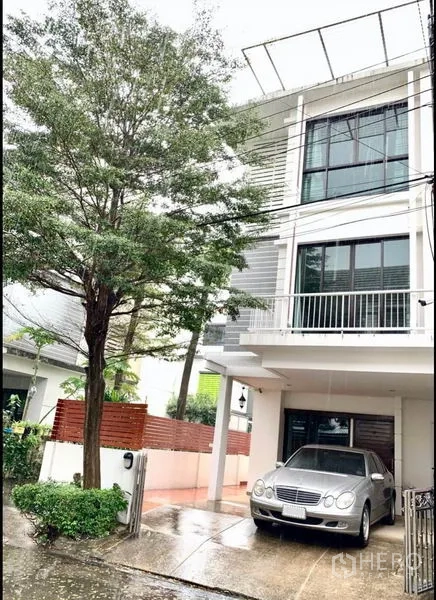 Townhouse for sale in Khlong Toei, Bangkok - Modern three-storey townhouse with balcony, large windows and a covered carport on a tree-lined lane.