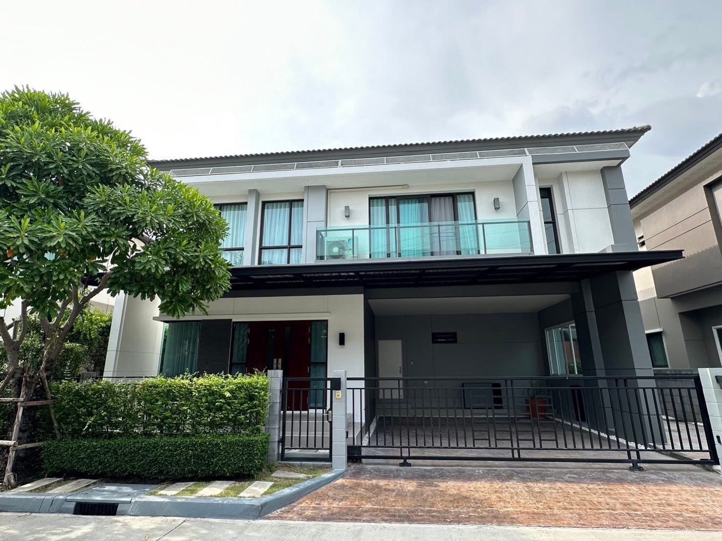 บ้าน ให้เช่าในBang Phli, Samut Prakan - ด้านหน้าบ้านเดี่ยวสองชั้นร่วมสมัย ระเบียงกระจก โรงจอดรถมีหลังคาและรั้วพุ่มไม้