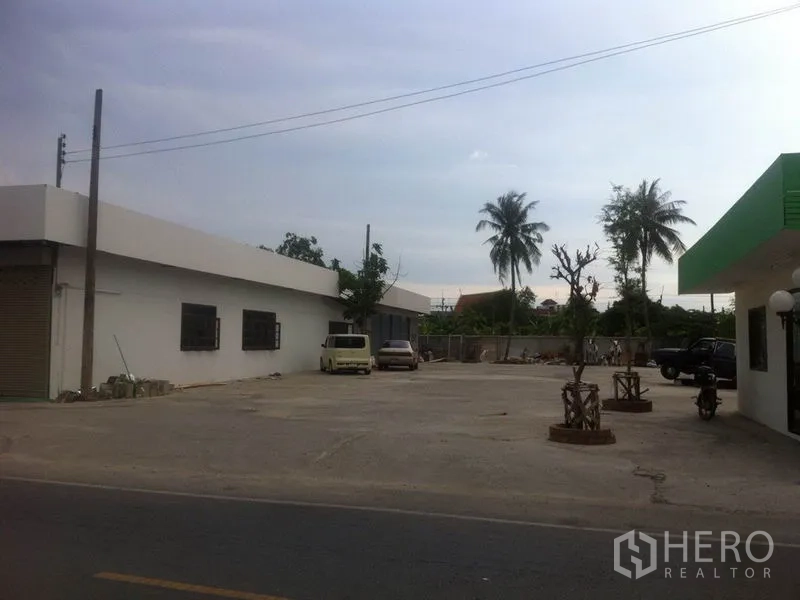 Warehouse for rent in Lam Luk Ka, Pathum Thani - Roadside view of the warehouse frontage with a large paved yard and palm trees, ideal for truck access.