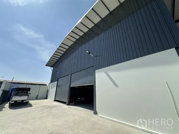 Warehouse for rent in Prawet, Bangkok - Side angle of the warehouse exterior showing two large roller doors and a broad driveway for trucks.
