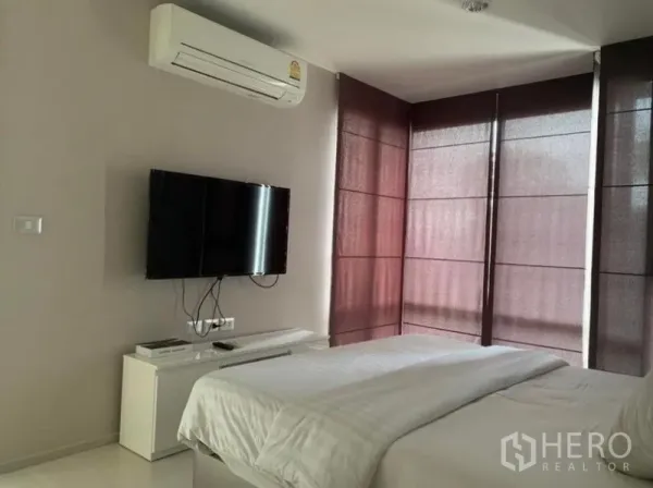 Condominium for rent in Khlong Toei, Bangkok - Bedroom view showing mounted TV, air conditioner, and corner windows with dark blinds.