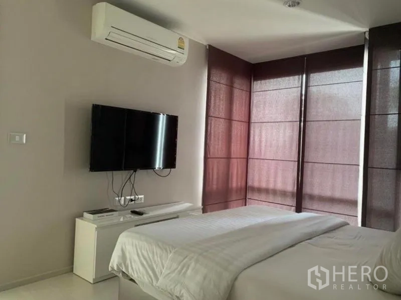 Condominium for rent in Khlong Toei, Bangkok - Bedroom view showing mounted TV, air conditioner, and corner windows with dark blinds.