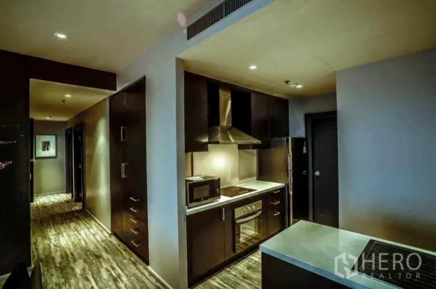 Condominium for rent or sale in Khlong Toei, Bangkok - Modern galley kitchen with built-in oven, cooktop, range hood and dark cabinetry along a corridor.