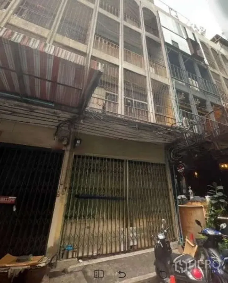 Shophouse for rent in Khlong Toei, Bangkok - Four-storey shophouse facade with metal folding gate and barred balconies on a narrow lane.