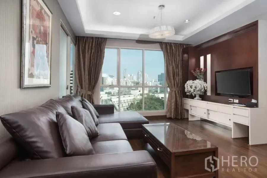 Condominium for rent or sale in Pathum Wan, Bangkok - Bright living room with leather sectional, TV wall, and large windows showcasing a Bangkok city view.