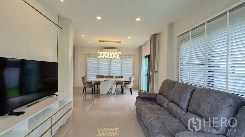 Detached House for rent in Samut Prakan - Open-plan living and dining area with sofa, TV wall and marble dining table.