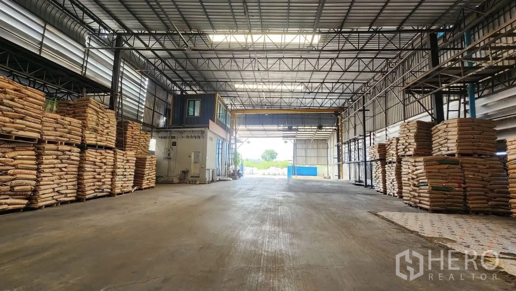 Warehouse for rent in Bang Phli, Samut Prakan - Interior of a high-clearance warehouse with steel trusses, skylights, stacked pallets, and a small office near a wide doorway.