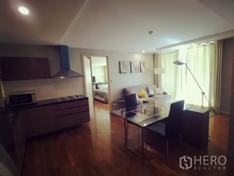 Condominium for rent or sale in Khlong Toei, Bangkok - Open-plan living and dining area with glass table, sofa and a modern kitchenette beside large curtained windows.