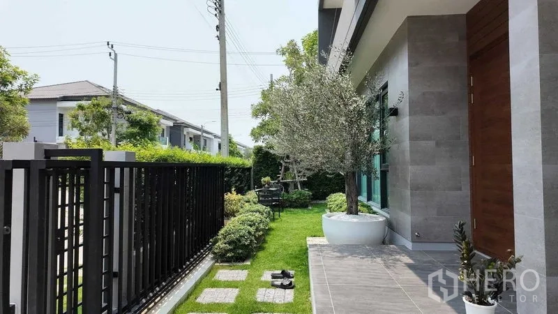 独立式住宅 出租或出售在Bang Na, Bangkok - 前门露台与修剪草坪，黑色金属栅栏旁的盆栽树。