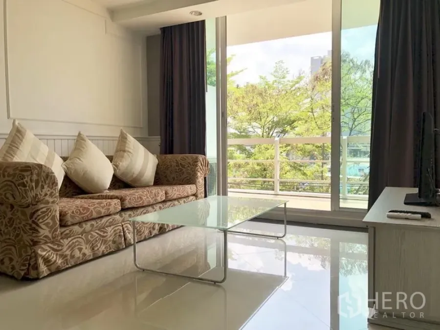 Condominium for rent in Khlong Toei, Bangkok - Living room opening to a balcony with leafy views through floor-to-ceiling sliding doors.