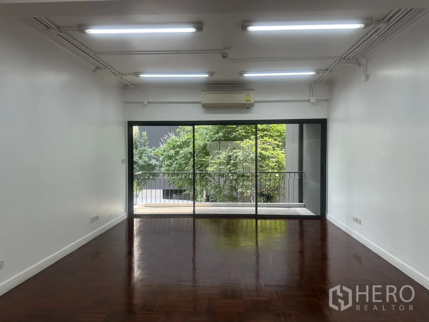 Office Space for rent in Watthana, Bangkok - Open-plan office with parquet floors, strip lighting, and sliding glass doors to a balcony with greenery view.
