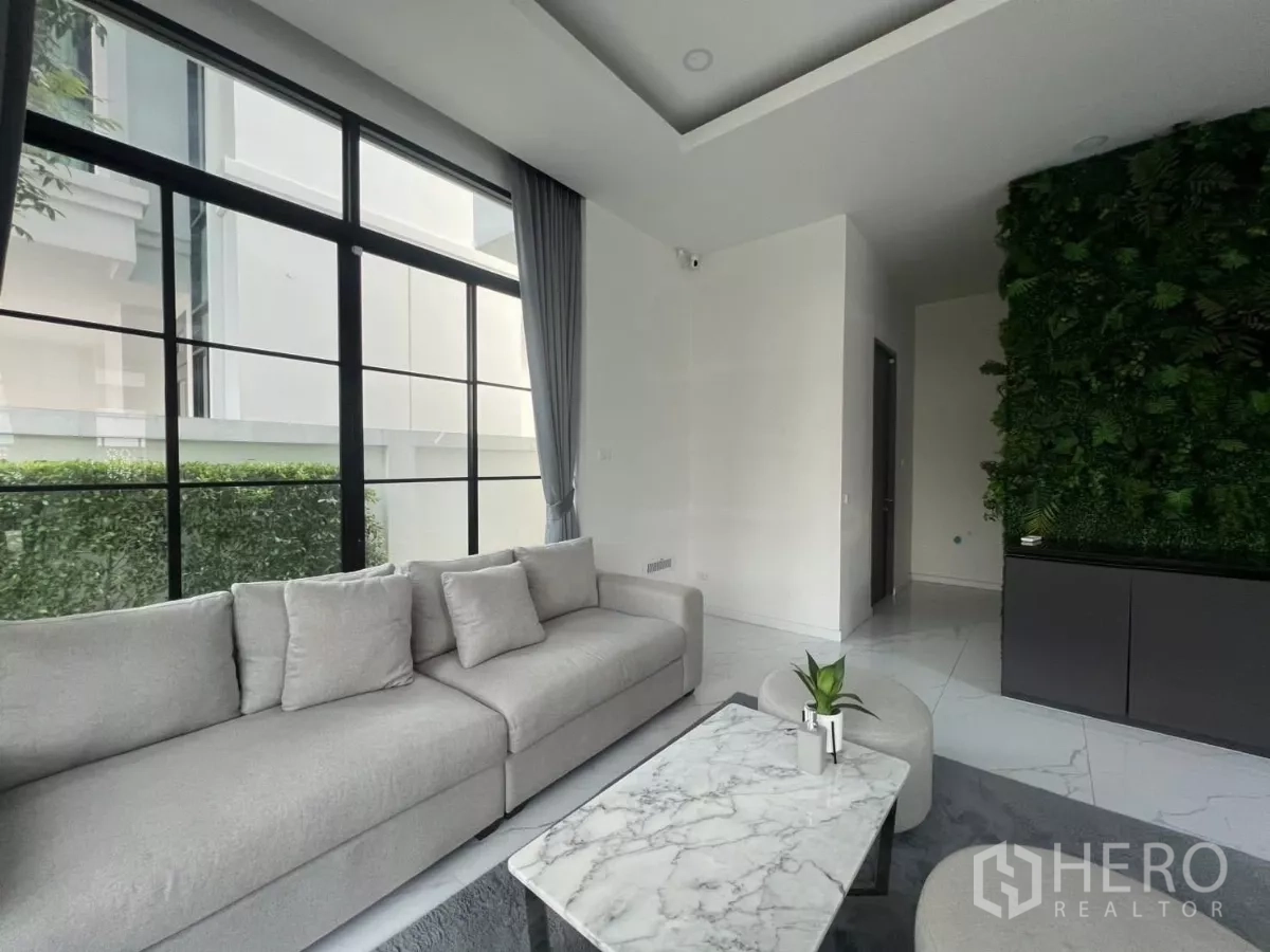Detached House for rent in Lat Phrao, Bangkok - Contemporary lounge with grey sofa, marble coffee table and floor-to-ceiling black-framed windows beside a vertical garden wall.