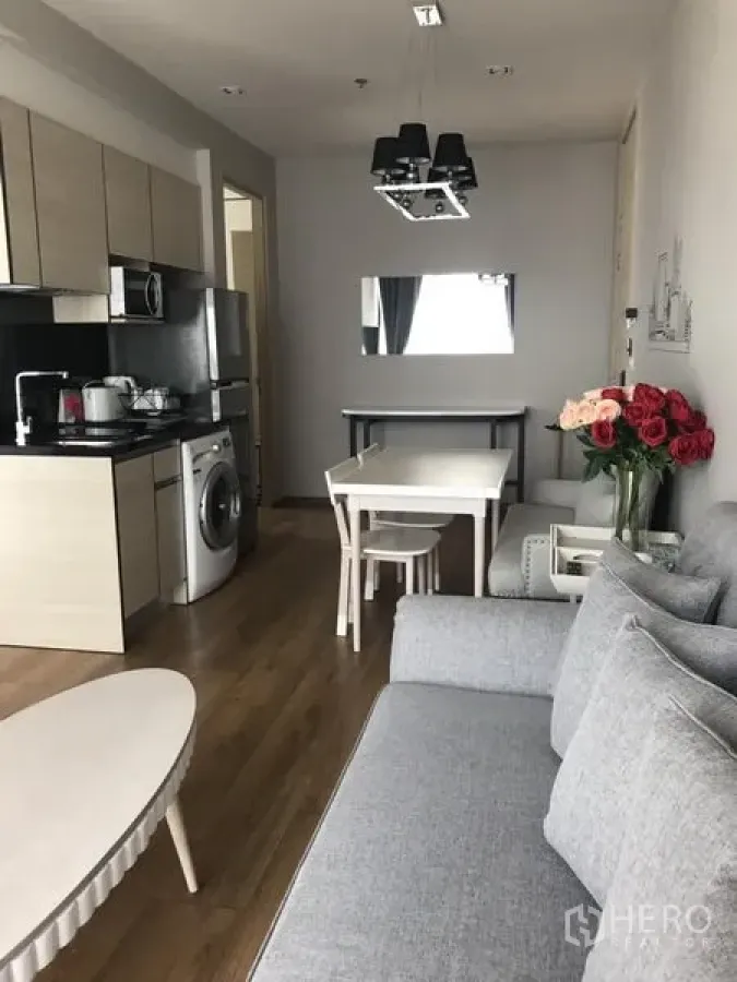 Condominium for rent in Khlong Toei, Bangkok - Open-plan living and dining area with kitchenette, wood floors, gray sofa and in-unit washing machine in a Sukhumvit condo.