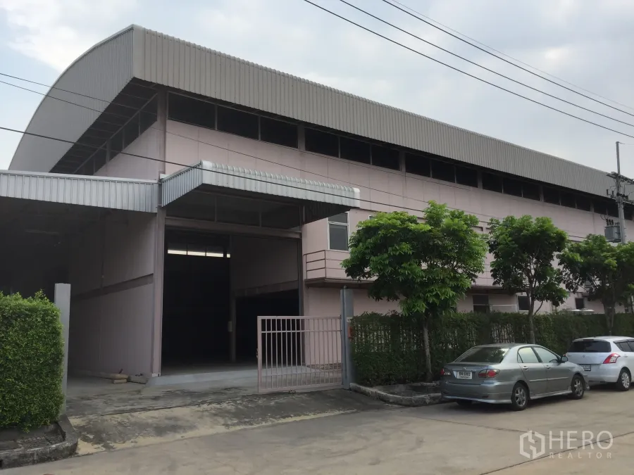 Factory for rent in Lam Luk Ka, Pathum Thani - Large factory exterior in Lam Luk Ka with roller shutter, gated entrance, trees and parking bays.