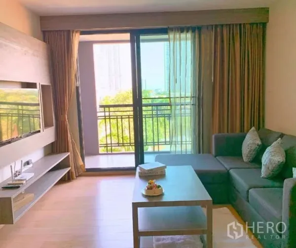 Condominium for rent - Bright living room with L-shaped sofa, TV wall, and balcony overlooking greenery.