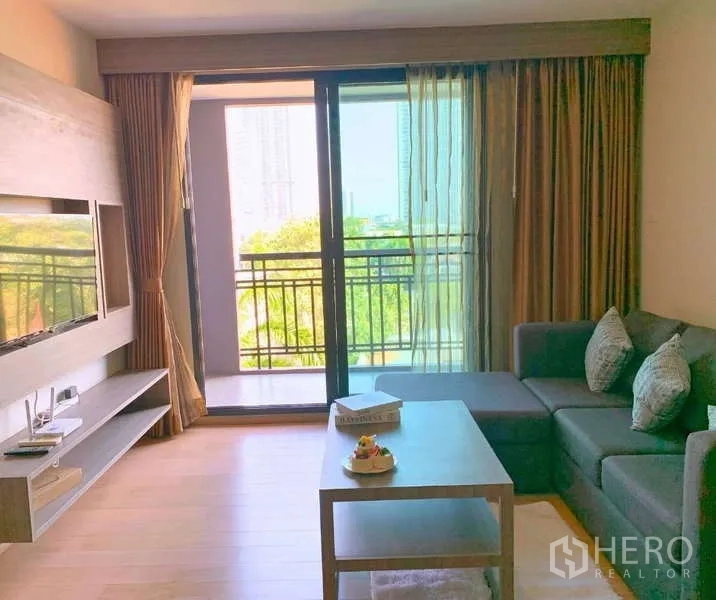 Condominium for rent - Bright living room with L-shaped sofa, TV wall, and balcony overlooking greenery.